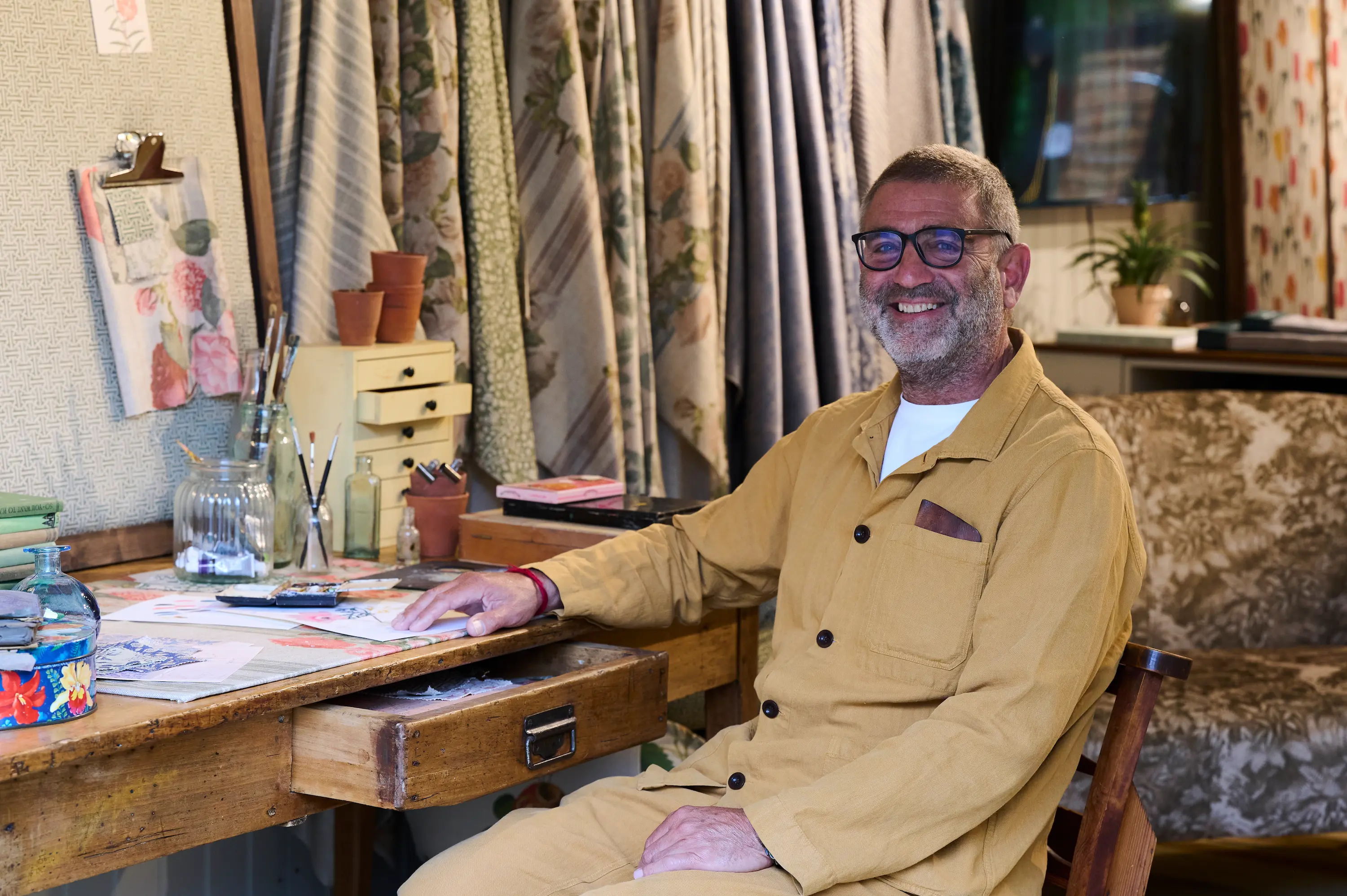 Founder Andrew Cussins seated at a worktable in a recreated artist’s studio, surrounded by botanical sketches, paintbrushes and floral fabrics at the RHS Chelsea Flower Show stand.